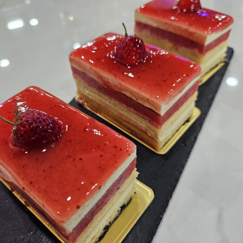 Trois parts de gâteau rectangulaires sur un plateau noir. Chaque part est recouverte d'un glaçage rouge brillant et décorée d'une fraise.