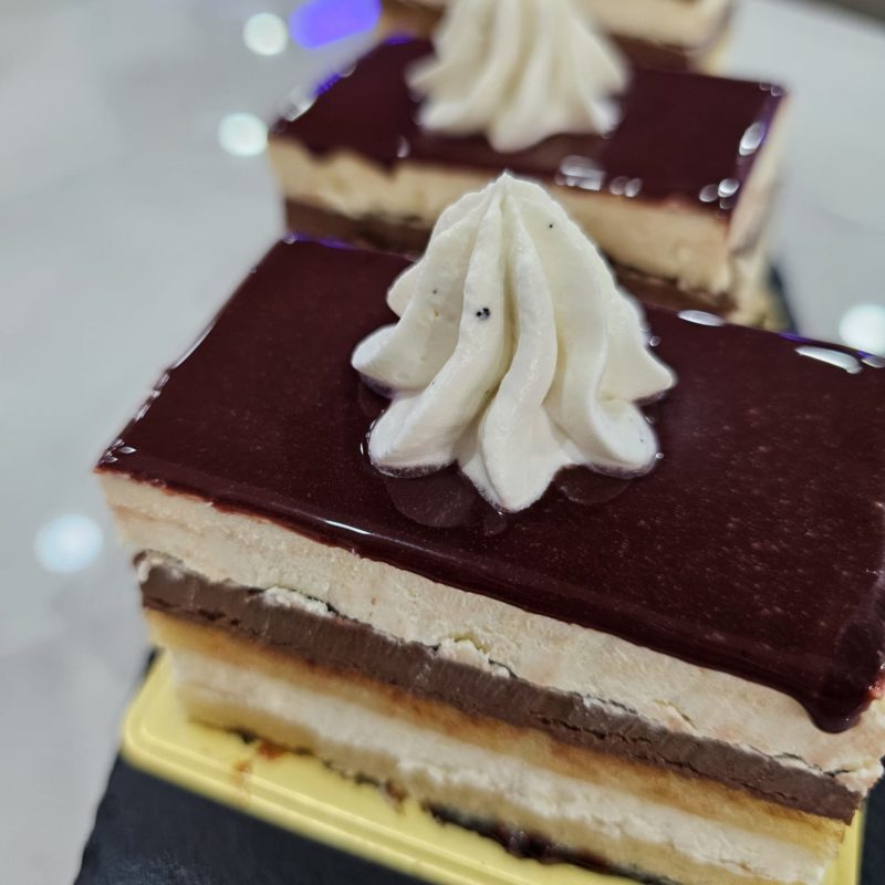 Trois parts de gâteau rectangulaires sur un plateau noir. Chaque part est recouverte d'un glaçage au chocolat et décorée d'une pointe de chantilly.