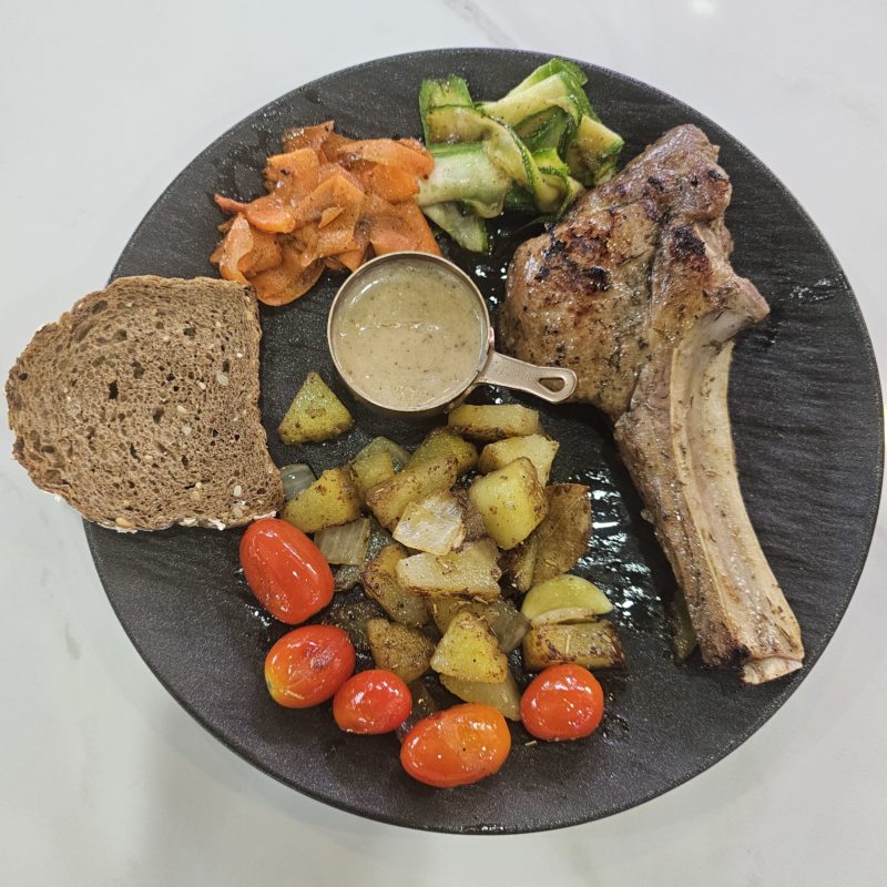 Une côte de veau grillée, présentée sur une assiette noire avec des pommes de terre rôties, des légumes grillés et une sauce crémeuse.