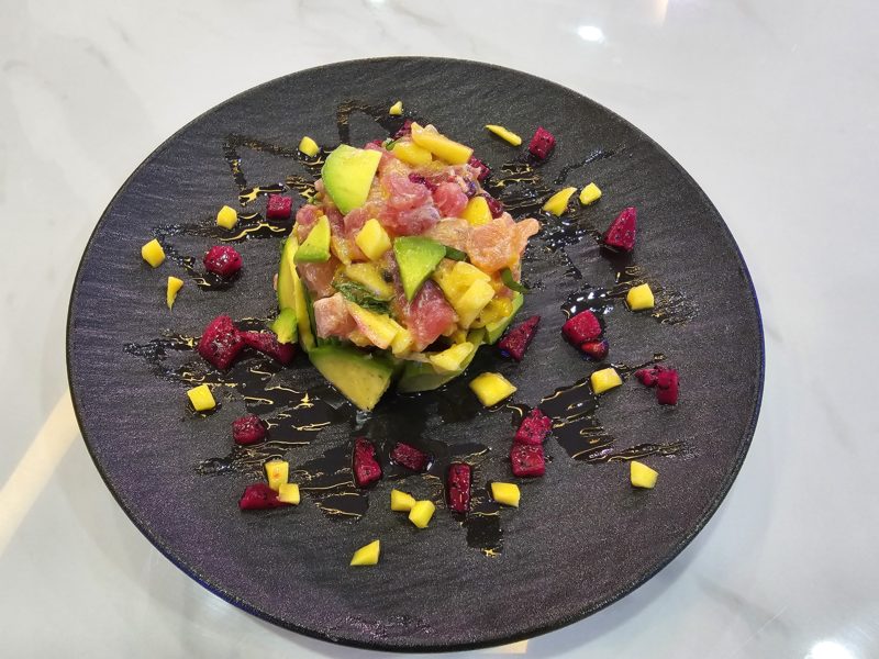 Un tartare de poisson cru composé de thon, de saumon, d'avocat, de mangue et de fruit du dragon, présenté sur une assiette noire.