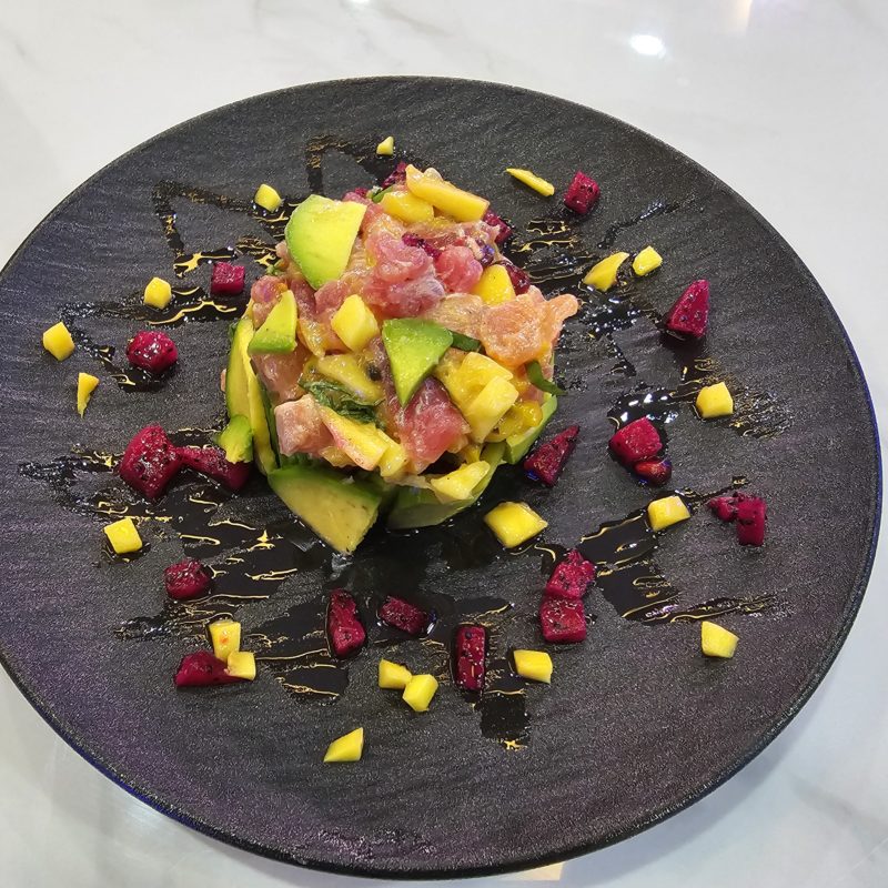 Un tartare de poisson cru composé de thon, de saumon, d'avocat, de mangue et de fruit du dragon, présenté sur une assiette noire.