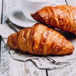 Deux croissants au beurre dorés posés sur une nappe en lin, à côté d'une tasse de café. Deux croissants au beurre dorés posés sur une nappe en lin, à côté d'une tasse de café.