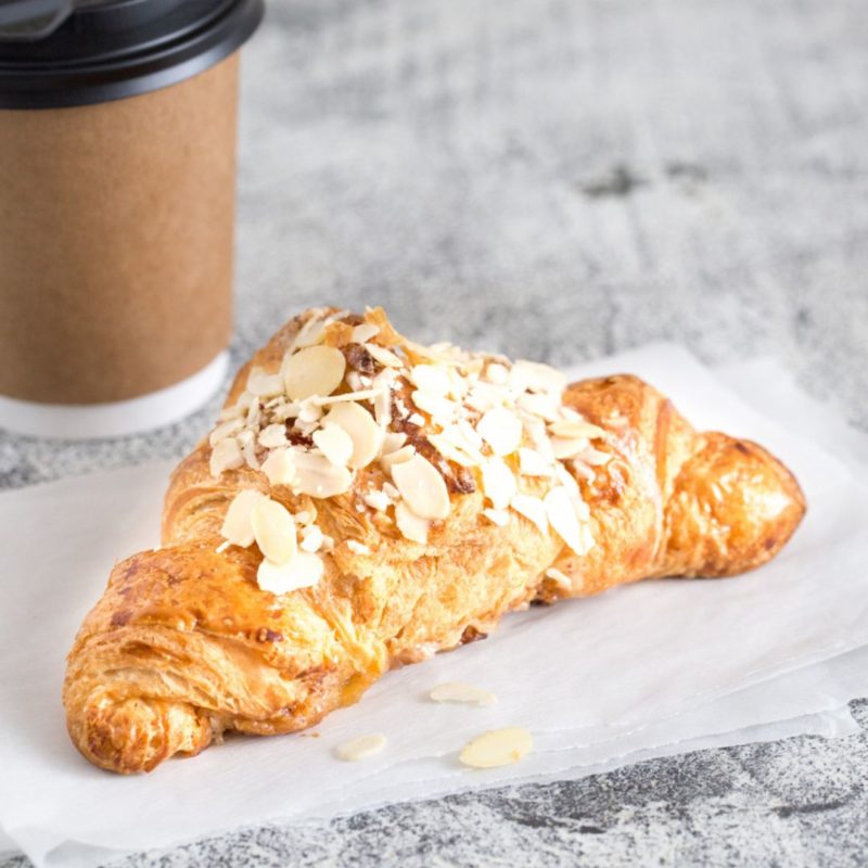 Un croissant aux amandes fraîchement sorti du four, posé sur du papier sulfurisé. Des amandes effilées sont éparpillées autour du croissant. Un gobelet à café en carton est visible en arrière-plan.