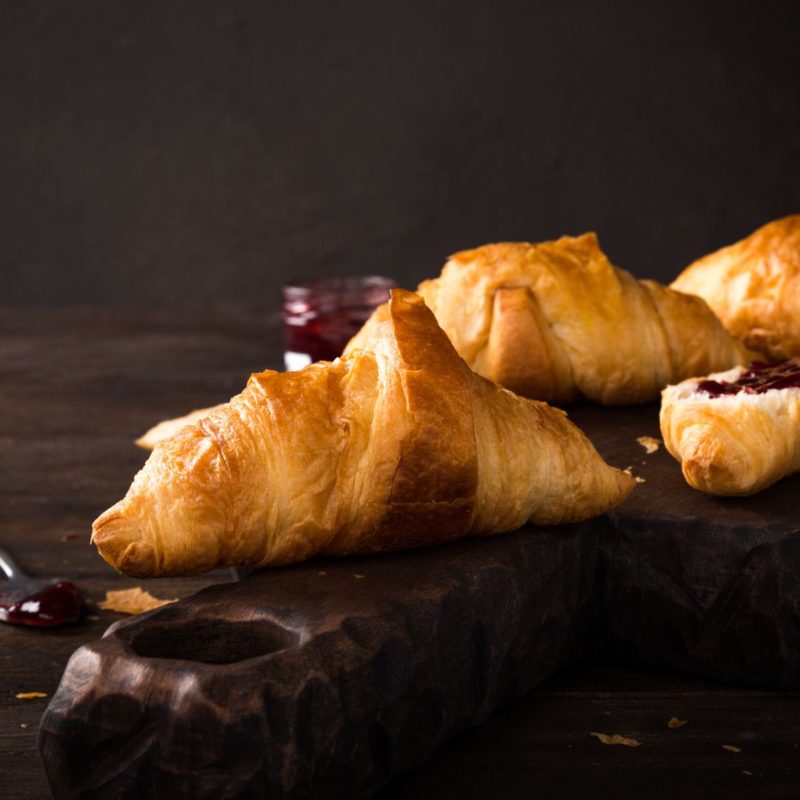 Quelques mini croissants dorés disposés sur une planche en bois. Un pot de confiture est visible en arrière-plan.