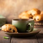 Une tasse de thé accompagnée d'un mini pain au chocolat sur une table en bois. D'autres mini pains au chocolat sont visibles en arrière-plan.