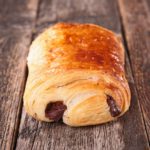 Un pain au chocolat doré posé sur une surface en bois. La pâte feuilletée est croustillante et le cœur de chocolat est visible.