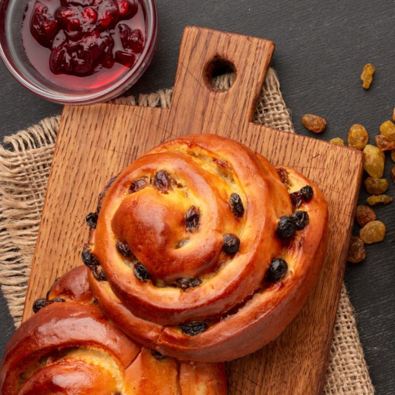 Deux pains aux raisins dorés posés sur une planche en bois. Un bol de confiture de canneberges et des raisins secs sont visibles à côté.