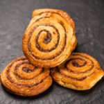 Une pile de roulés à la cannelle dorés, disposés sur une surface noire. Chaque roulé est parsemé de sucre et présente des couches de pâte feuilletée.