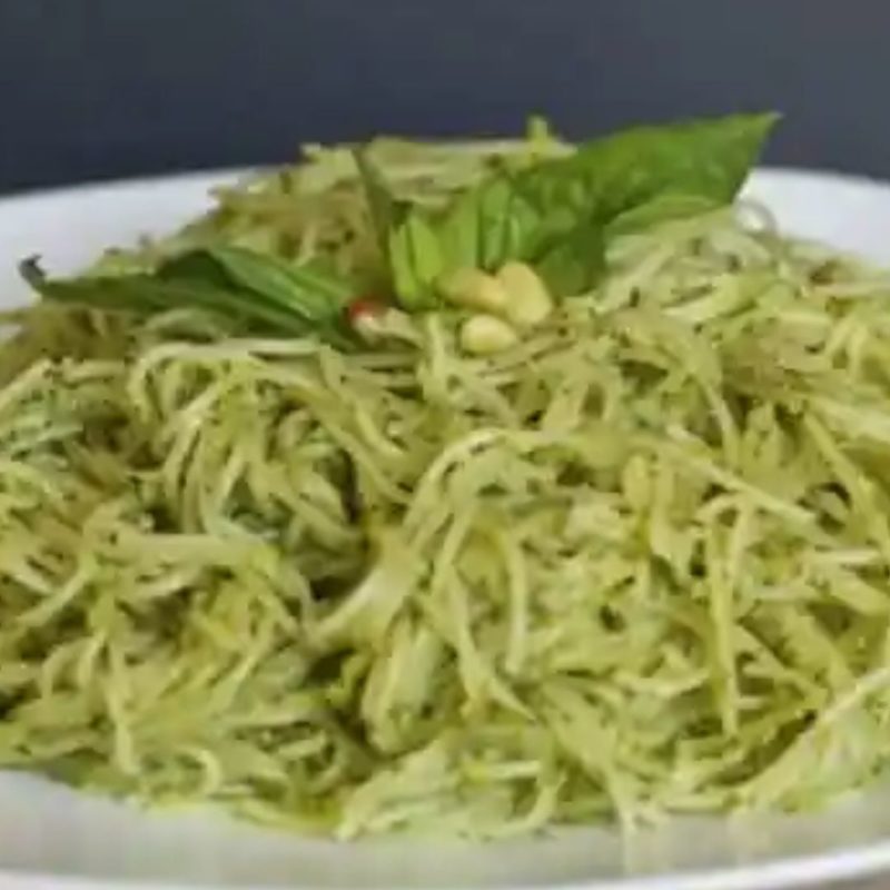 Un plat de spaghetti enroulés dans une sauce pesto verte brillante, garnis de quelques feuilles de basilic frais et de pignons de pin. Des morceaux de poulet grillé sont éparpillés sur le dessus.